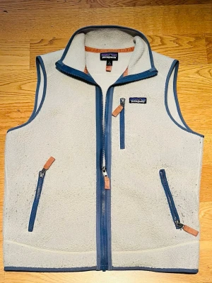 Beige fleeceväst från Patagonia - Mysig beige fleeceväst från Patagonia med blå detaljer och dragkedjor. Västen har tre fickor med dragkedja och hög krage. Perfekt för lager-på-lager och outdoor-stil. Tillverkad i mjukt fleece-material och har snygga läderdetaljer på dragkedjorna.