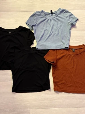 4-pack croppade ribbade toppar - Säljer fyra croppade toppar i svart, ljusblå och rostbrun färg. Alla har rund halsringning och korta ärmar, perfekta till jeans eller kjol. De är ribbade och sitter snyggt slim på kroppen.