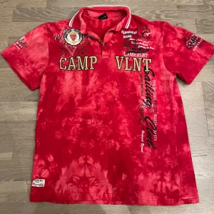 Röd t-shirt Camp VLNT  - Säljer en röd tie-dye t-shirt från Camp VLNT med broderade och tryckta detaljer, text och emblem på bröstet och ärmarna. T-shirten har krage med vita ränder och korta ärmar. Perfekt för dig som gillar unika och färgstarka plagg. Tröjan är 4xl men sitter som L. Tröjan knappast använd och skit fet👍