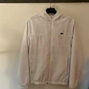 Vit vindjacka från Lacoste Sport XS - Fräsch vit vindjacka från Lacoste i storlek Xs. Jag säljer nu min Lacoste jacka eftersom den inte passar mig längre då jag köpte den för 1950 i butik. Jackan ser helt ny ut och har en dragkedja hela vägen, luva och elastiska muddar. Klassisk krokodillogga på bröstet och två fickor framtill. Perfekt för dig som gillar clean och sportig stil.