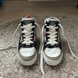 Svarta och vita sneakers, Aldrig använda! - Snygga sneakers i svart, vitt och grått med coola detaljer och orange flärp bak. Skorna har snörning med röda öglor och en chunky vit sula. Materialet är syntet och designen är modern med kontrasterande paneler. Perfekta för dig som gillar streetstyle.