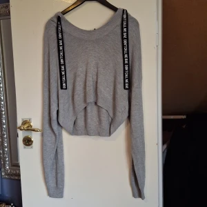 Grå croppad hoodie med textband - Grå croppad hoodie med långa ärmar och svarta band med texten 'DON'T CALL ME BAE' längs axlarna. Tröjan är stickad och har en oversized passform med ribbade muddar och huva. Perfekt för en chill och trendig look.