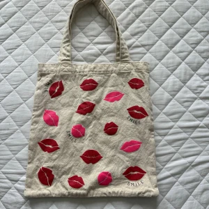 Beige väska med läppar  - Superfin beige shoppingväska i 100% bomull från Kappahl, dekorerad med röda och rosa läppar samt texten 'SMILE'. Perfekt storlek för att bära med sig allt man behöver och har två rejäla handtag. Väskan är både söt och trendig.