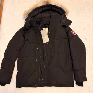 Svart dunjacka från Canada Goose - Svart dunjacka från Canada Goose med ikonisk röd logotyp på ärmen. Jackan har en fluffig beige päls runt huvan, dragkedja och knappar framtill. Perfekt för kalla dagar med sin tjocka och värmande design. Snygg och klassisk look med lång ärm och normal passform.