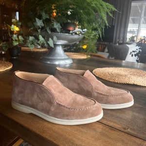 Bruna mockaskor från Loro Piana - Säljer ett par stilrena bruna mockaskor från Loro Piana med vit platt sula. Skorna har en minimalistisk design utan snörning och är tillverkade i mjuk mocka för en lyxig känsla. Perfekta för dig som gillar klassisk och clean stil. Skorna är i toppskick, skriv vid frågor eller funderingar!