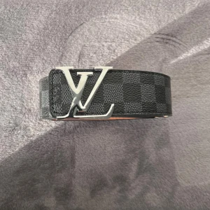 Lv bälte  - Säljer ett Louis Vuitton bälte i svart och grått med rutigt mönster och en stor LV-logga i silver på spännet. Bältet är tillverkat i skinn och har detaljerad sömnad längs kanterna. Insidan är ljusbrun med präglad Louis Vuitton-text.