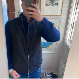 Svart väst från Oscar Jacobson - Snygg svart väst från Oscar Jacobson med hög krage och hel dragkedja framtill. Västen har en stilren design med diskreta fickor på sidorna och lätt quiltade detaljer på ryggen. Perfekt att slänga över en hoodie eller tröja när det är kyligt ute.