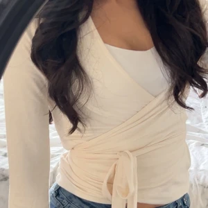 Vit omlottopp med knytband - Säljer en fräsch vit omlottopp med långa ärmar och snygg knytning i midjan. Toppen har en v-ringad design och är gjord i mjukt bomullsmaterial. Perfekt att styla med jeans för en clean och trendig look.