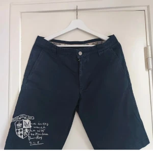 La Martina Shorts M - Snygga mörkblå shorts från La Martina med broderad logga och text på ena benet. Klassisk passform med knapp och dragkedja framtill samt sidofickor. Perfekta för sommardagar när du vill ha en stilren look.