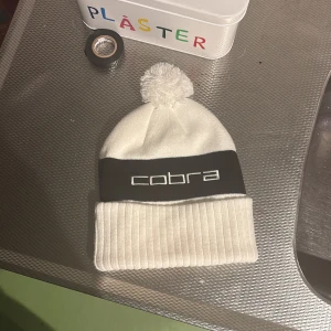 Vit och svart mössa med tofs - Snygg vit mössa från Cobra med svart band och broderad logga framtill. Mössan har en stor tofs på toppen och bred ribbad uppvikning. Perfekt för kalla dagar och ger en stilren look.