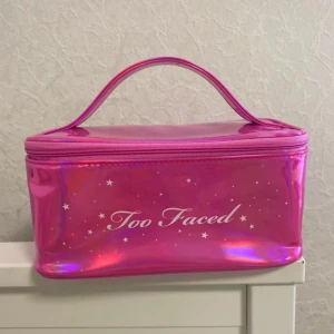 Rosa necessär från Too Faced - Superfin rosa necessär från Too Faced med holografisk finish och vita stjärndetaljer. Väskan har ett praktiskt handtag upptill och stängs med dragkedja. Perfekt för smink eller småprylar, och den är tillverkad i glansig syntet.