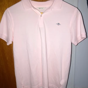 Ljusrosa pikétröja från GANT - Snygg ljusrosa pikétröja från GANT med klassisk krage och korta ärmar. Tröjan har broderad GANT-logga på bröstet och är gjord i mjuk bomull. Perfekt för en clean och fräsch look.