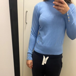 Ljusblå polotröja med knappar - Snygg ljusblå polotröja med ribbad krage och muddar. Tröjan har långa ärmar och tre dekorativa knappar i guld på vardera ärmslut. Materialet känns mjukt och är troligtvis bomull eller en syntetblandning. Perfekt för dig som gillar stilrena och klassiska plagg.