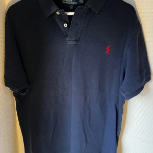 Marinblå pikétröja från Ralph Lauren - Klassisk mörkblå pikétröja med kort ärm och röd broderad logotyp på bröstet. Slim fit modell så den passar L. 