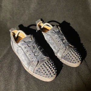 Svarta Louboutin sneakers med nitar - Säljer ett par svarta sneakers från Christian Louboutin i mocka med ikoniska röda sulor. Skorna har snörning och coola svarta nitar på tån för en edgy vibe. Insidan är ljus och sulan har Louboutin-loggan. Perfekta för dig som vill sticka ut. ÄKTA 