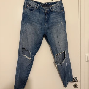 Blå ripped jeans från ONLY (kort modell) - Snygga blå jeans från ONLY med slitna detaljer och stora hål vid knäna. Klassisk femficksmodell med normal passform och dragkedja. Perfekta för en avslappnad streetstil. Jeansen är i ett mjukt denimtyg och har en något avsmalnande benform.