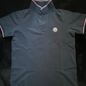 Mörkblå Moncler pikétröja - Snygg mörkblå pikétröja från Moncler med klassisk krage och knappar framtill. Tröjan har Moncler-logga på bröstet och diskreta rödvita detaljer på krage och ärmslut. Perfekt för dig som gillar stilrena och exklusiva plagg.