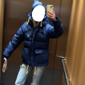 Mörkblå dunjacka från Fjällräven - Säljer en mörkblå dunjacka från Fjällräven med gul snörning vid huvan och nederkant. Jackan har en puffer-design, två stora fickor med knappar och dragkedja framtill. Perfekt för kalla dagar. Jackan är i storlek S och har inga defekter!