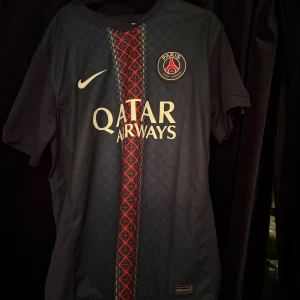 PSG matchtröja Nike blå/röd - Snygg Paris Saint-Germain fotbollströja från Nike i mörkblått med röd och vit grafisk rand mitt på bröstet. PSG-logga och Nike swoosh på bröstet samt Qatar Airways-tryck. Klassisk passform och korta ärmar, perfekt för fotboll eller supporterliv.