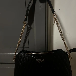 Svart axelväska från Guess - Snygg svart axelväska från Guess med guldfärgade kedjedetaljer. Väskan har dragkedja upptill och extra fack med dragkedja på baksidan.