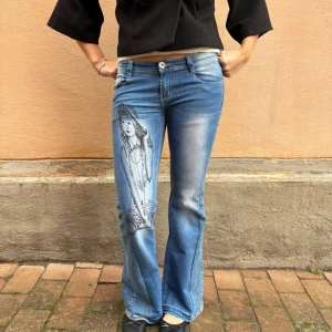 002 jeans med motiv  - Ett par perfekt lågmidjade jeans i en mellanblå tvätt. Dem har ett svart motiv på höger men med silver detaljer. Två insydda kilar på sidan av varje byxben. Fickorna har en unik design med knapp som gör att dessa jeansen är unika och kan göra en tråkig outfit snyggare. Modellen är 175cm och de passar bra i längden. Det står i jeansen att den är storlek 40 men liknar mer storlek 36/38. Midjemått: 85cm. Innerbens längd: 85cm