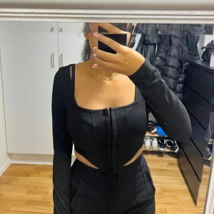 Svart långärmad korsett-topp - Säljer en svart korsett-topp med lång ärm och fyrkantig urringning. Toppen har en snygg croppad passform med dragkedja framtill och coola cut-outs vid midjan. Perfekt för dig som gillar trendiga och edgy plagg.