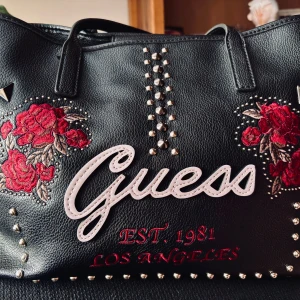 Snygg vintageväska från Guess!  - Snygg vintageväska i konstläder med stor kapacitet från Guess med broderade rosor och nitar. Mått: 45 x 25 cm. Köpt på second hand i USA för 10 år sen. I perfekt skick! 