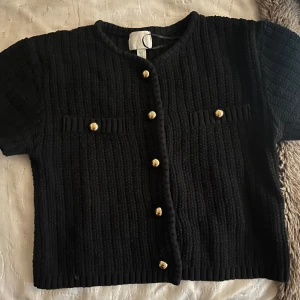 Svart stickad bolerotopp med guldfärgade knappar - Supersnygg svart stickad bolerotopp från H&M med korta ärmar och croppad passform. Toppen har guldfärgade knappar framtill och dekorativa fickor på bröstet. Perfekt för dig som gillar en chic och trendig look med lite edge.