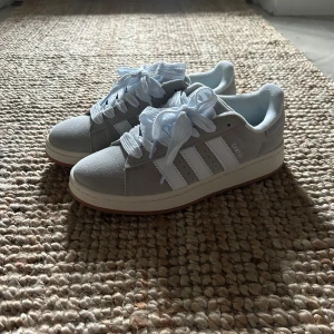 Adidas Campus sneakers ljusgrå - Säljer ett par Adidas Campus sneakers i ljusgrå mocka med vita detaljer och klassiska tre ränder på sidan. Skorna har vit platt sula, rund tå och breda vita skosnören. Perfekt för dig som gillar stilrena och tidlösa sneakers.