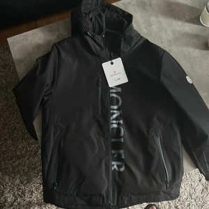 Svart vindjacka från Moncler med huva - Svart vindjacka från Moncler med stor logga längs framsidan, huva och tre dragkedjeförsedda fickor. Jackan har lång ärm och Moncler-märke på ena armen. Snygg och clean design, perfekt för dig som gillar streetwear.