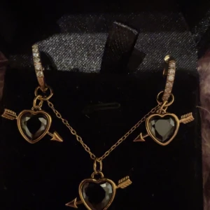 Guldörhängen och halsband med svarta hjärtan guld  - Säljer ett set med örhängen och halsband i guldton med svarta hjärtformade stenar och pil-detaljer. Örhängena har glittriga stenar på bågen och hängande hjärtan med pil. Halsbandet matchar med likadant hjärthänge. Perfekt för dig som gillar unika smycken med cool design. Rostfria