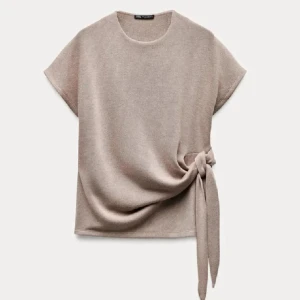 Beige topp med knyt från Zara - Säljer en stilren beige topp från Zara med rund hals och korta ärmar. Toppen har en snygg knytning i sidan som ger en cool detalj och lite draperad effekt. Perfekt för dig som gillar enkel men trendig design. I super bra skick!!storlek S