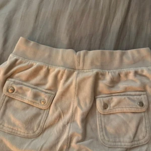 Beiga mjukisbyxor Juicy Couture S - Supermysiga beiga mjukisbyxor från Juicy Couture i velour med snörning i midjan, fickor fram och bak med lock och knappar samt broderad JC-logga. Perfekta för chill dagar och har en klassisk loose passform. Knappen är borta från ena fickan men inget som syns när man har de på sig. 