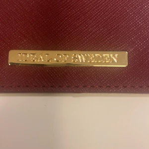 Vinröd plånbok från iDeal of Sweden - Slimmad vinröd plånbok från iDeal of Sweden med guldfärgad metallbricka framtill. Plånboken har flera kortfack och är tillverkad i ett strukturerat syntetmaterial som ger en lyxig känsla. Perfekt för dig som vill ha stilren och smidig förvaring av kort. Oanvänd 