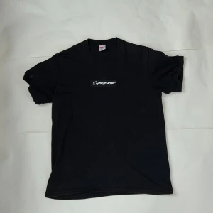 Svart Supreme t-shirt med tryck - Svart t-shirt från Supreme med vit logga framtill och texten 'You was raised off our shit' i vitt på ryggen. Klassisk passform och rund hals. Tillverkad i mjuk bomull, perfekt för en streetwear-look.