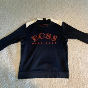 Marinblå tröja från Hugo Boss - Snygg marinblå tröja från Hugo Boss med vita axelpartier och röd logga framtill. Tröjan har rund hals, långa ärmar och är tillverkad i mjuk bomull. Perfekt för dig som gillar stilrena och sportiga plagg med premiumkänsla.