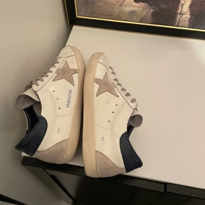 Golden Goose Superstar sneakers vit/grå - Golden Goose Superstar sneakers i vitt skinn med grå mockadetaljer och stjärna på sidan. Mörkblå häl och grå snörning ger en cool kontrast. Platt sula och rund tå. Perfekt för dig som gillar streetstyle och unika detaljer.