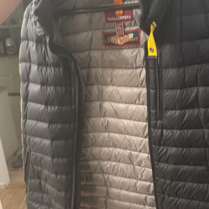 Svart dunväst från Parajumpers XL - Säljer en svart dunväst från Parajumpers i storlek XL. Västen är quiltad med horisontella sömmar, har en gul detalj vid kragen och dragkedja framtill. Insidan är ljusgrå och västen är lättviktig, perfekt för lager-på-lager-stil. Äkta och har kvitto köpts för 3500