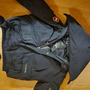 Svart dunjacka från Canada Goose - Svart dunjacka från Canada Goose med huva, ribbade muddar och flera fickor framtill. Jackan har klassisk logotyp på ärmen och broderad etikett på bröstet. Perfekt för kalla dagar med sin tjocka och isolerande design. Dragkedja och knappar framtill. Lämnad till kemtvätt 