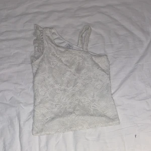Vit spets topp från Lindex - Söt vit topp från Lindex med blommig spets och asymmetrisk design. Ena axeln har en smal axelrem och den andra en volangdetalj. Perfekt för sommarens varma dagar eller när du vill ha något extra fint.