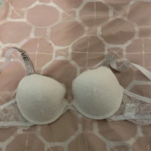 Vit spetsig bandeau-tubtopp med strass - Supersnygg vit bandeau-tubtopp med spets och vadderade kupor. Axelbanden är täckta av glittriga strass och har texten 'LOVE' i svarta stenar. Spetsdetaljer längs kanterna ger en extra lyxig vibe. Perfekt för dig som gillar att sticka ut med coola detaljer.