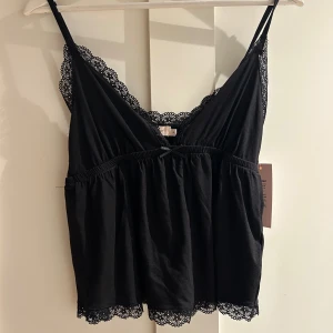 Svart spetskantad linnetopp från Nelly - Supersöt svart linnetopp från Nelly med tunna axelband, v-ringning och dekorativ spetskant längs halsringning och nederkant. Toppen har en liten rosett framtill och är lätt rynkad under bysten för en luftig känsla. Perfekt att styla med jeans eller kjol.