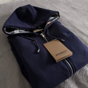 Burberry tröja  - Snygg marinblå hoodie från Burberry med klassisk rutmönstrad insida på huvan och broderad logga på bröstet. Tröjan har hel dragkedja, snörning i huvan och två fickor framtill. Perfekt för dig som vill ha en stilren och exklusiv look.