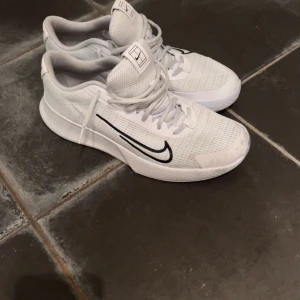 Nike Vapor Lite vita inomhusskor.✨ - Nike Vapor Lite sneakers i vitt med svart swoosh på sidan. Skorna har mesh-ovandel för bra ventilation, snörning och platt sula. Snygg sportig design med logga på hälen och tungan. Perfekta för dig som gillar stilrena och bekväma inomhusskor. | har använts till padel så de är lite blåa efter sanden på banan men det går att tvätta bort. | För mer frågor och funderingar skriv till mig.✨