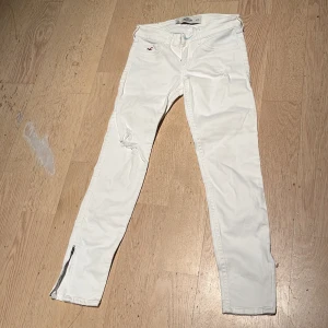 Vita skinny jeans från Hollister - Snygga vita skinny jeans från Hollister med dragkedjor vid bensluten och slitningar på båda benen. Klassisk femficksmodell med broderad logga och detaljerade bakfickor. Perfekta för dig som gillar en fräsch och stilren look. Pris kan diskuteras vid snabb affär
