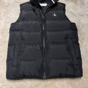 Svart dunväst från Calvin Klein - Stilren svart dunväst från Calvin Klein med broderad CK-logga på bröstet och dragkedja framtill. Insidan har kontrasterande vit panel med stor logotyp. Västen har hög krage och två sidofickor, perfekt för lager-på-lager under kyliga dagar.