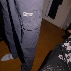 Grå cargobyxor från McKenzie - Säljer ett par grå cargobyxor från McKenzie med elastisk midja och stora fickor på sidorna. Byxorna har en avslappnad passform och är tillverkade i ett lätt syntetmaterial, perfekta för en chill och street-inspirerad stil. Finns även en jacka till den!!