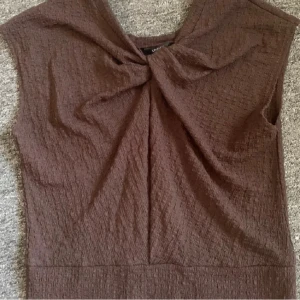 Brun ärmlös topp med twistad detalj - Säljer en brun ärmlös topp från Lindex med twistad detalj framtill vid halsen. Toppen har en lätt strukturerad yta och rund halsringning. Perfekt att styla med jeans eller kjol för en cool look. Aldrig använd!