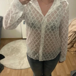 Vit transparent skjorta med mönster - Säljer en vit transparent skjorta med rutigt mönster och klassisk krage. Skjortan har långa ärmar och knäppning framtill. Perfekt att styla lager på lager för en trendig look.