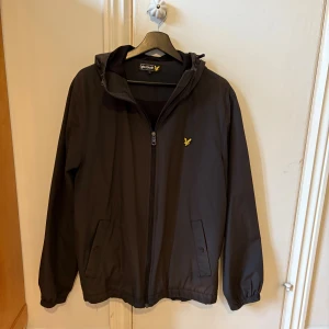 Vindjacka Lyle&Scott - Svart vindjacka med huva. Sidofickor med knapp. Lite ändringar i färg (svart/grå) vid linningen. 